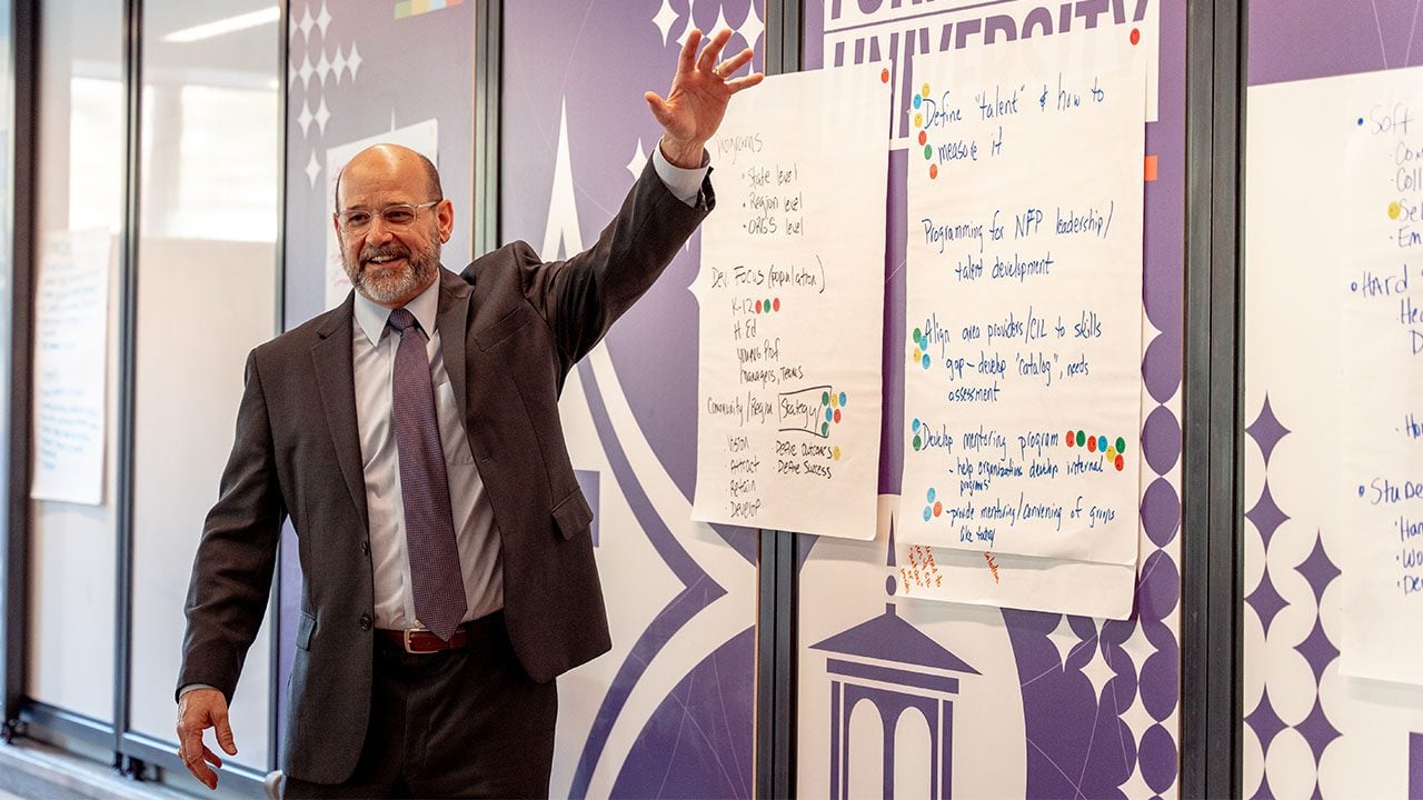 A man wearing a suit and tie gestures to a group off-camera as he presents in front of a number of handwritten signs with notes.