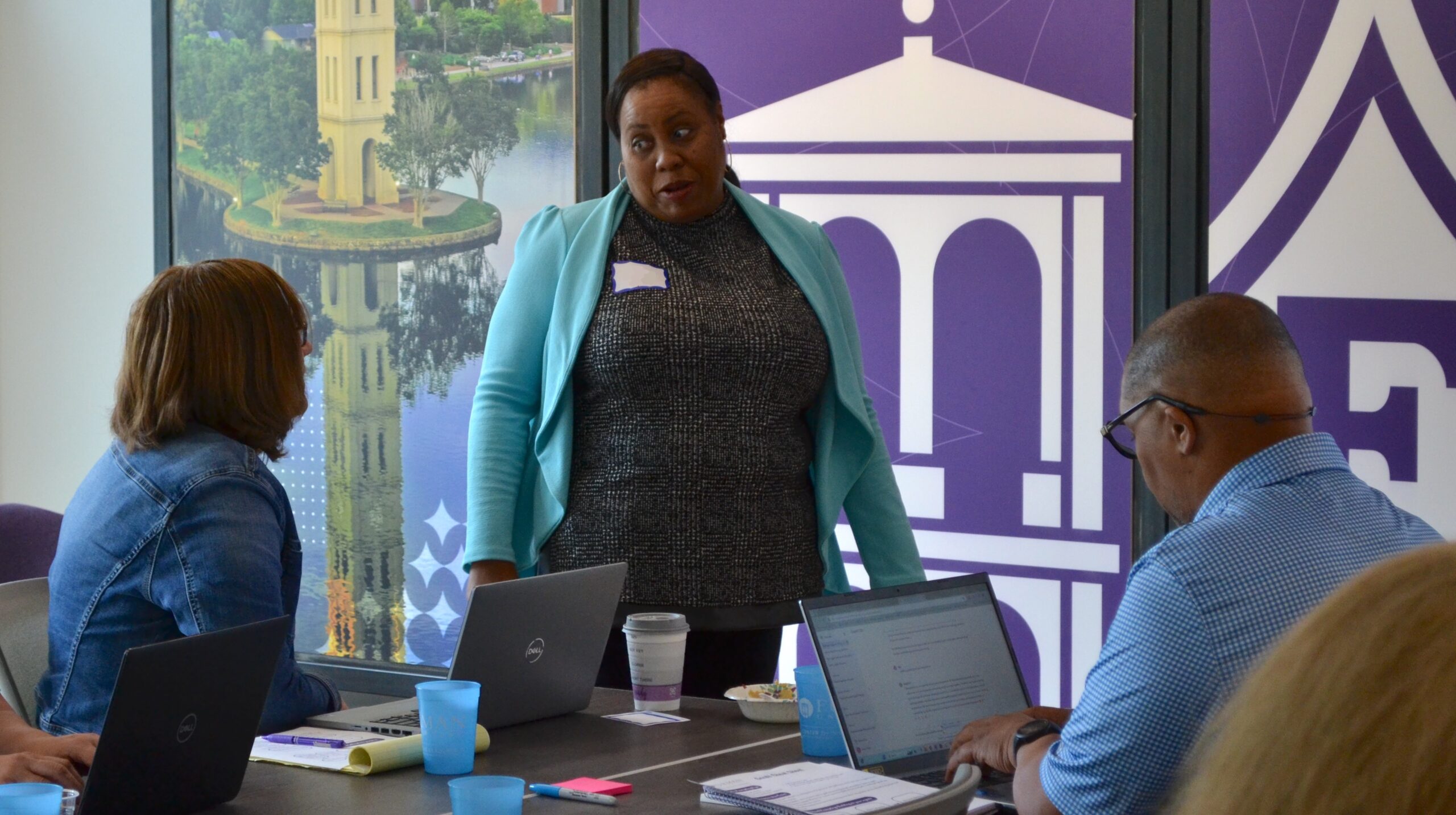 A woman in a blue blazer wears a nametag and speaks to two professionals sitting at a table in front of her.